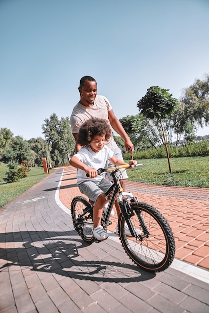 AfroAmerican man helping his curly kid with learning to ride a bicycle