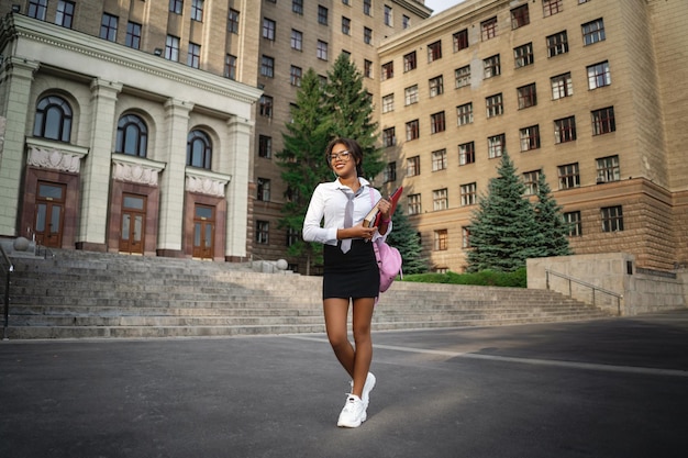 Foto studentessa afroamericana con zaino in piedi e con in mano libri vicino al vecchio edificio maestoso