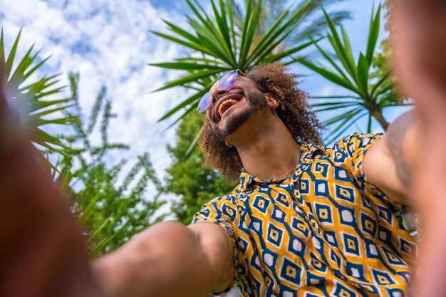 ビーチの隣のいくつかのヤシの木の隣で夏休み中のアフロヘアーの男が両手で笑顔で自分撮りをする旅行と観光のコンセプト