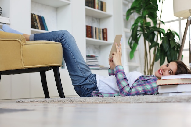 Afstand leren. Jonge vrouwelijke student die thuis studeert, artikel schrijft voor publicatie, zittend op de vloer met tablet tegen een gezellig interieur, omringd met stapel boeken