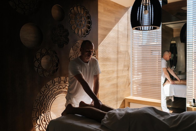 Afstandsbeeld van een mannelijke masseur die een essentiële olie massage geeft aan de kalfspieren van een onherkenbare man die op een massage tafel ligt in een donkere kamer of in een luxe spa salon bij het raam Concept van professionele massage