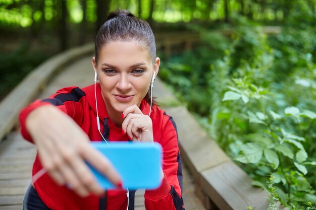 Afstandsonderwijs concept. Een aantrekkelijk meisje in sportkleding zit op een houten pad in een bospark. Online lessen via smartphone.