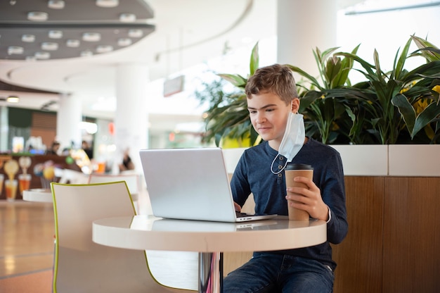 Afstandsonderwijs een tiener met een medisch masker zit aan een tafel in een café met een laptop en een mok...