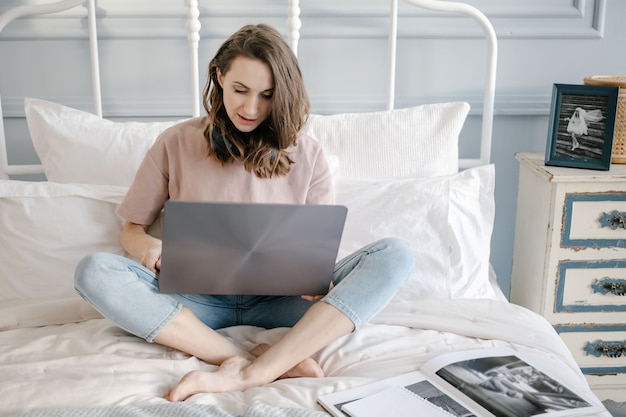 Afstandsonderwijs, online onderwijs en werk. Gelukkige vrouw in casual met de koptelefoon bezig met een laptop op afstand van huis op bed.