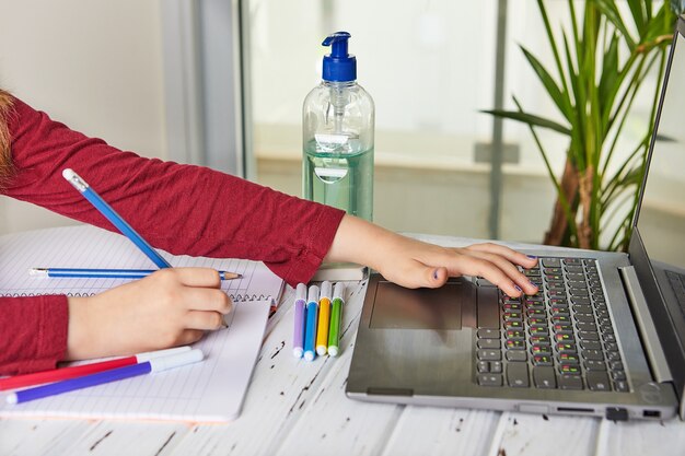 Afstandsonderwijs online onderwijs. Schoolmeisje dat thuis met laptop bestudeert en schoolthuiswerk doet. Trainingsboeken en gekleurde viltstiften op tafel, gel met 70 procent alcohol