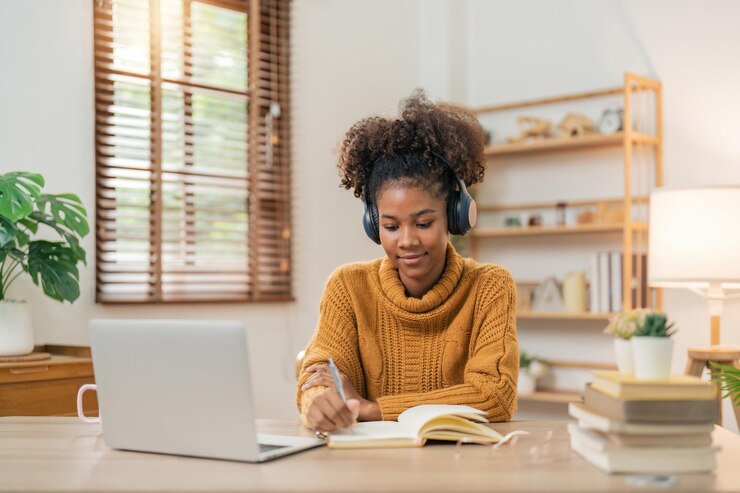 Foto afstandsonderwijs portret van lachende afro-amerikaanse vrouw die aan het bureau zit met behulp van een laptop en in een notitieboekje schrijft en aantekeningen maakt, kijkt naar een tutoriallezing of webinar en thuis online studeert