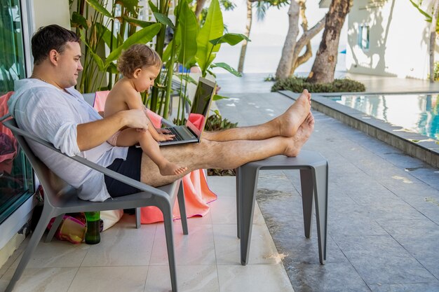 afstandswerk. man met een peuter in zijn armen met een laptop bij het zwembad in een tropisch land