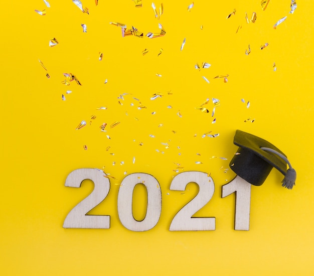 Afstuderen 2021. Houten nummer 2021 in een afgestudeerde hoed met confetti op een gele achtergrond bovenaanzicht.