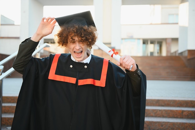 Afstuderen aan de universiteit Jonge glimlachende jongen universitair afgestudeerd in traditionele bonet en mantel die staat en diploma in de hand houdt over de achtergrond van het universitaire gebouw