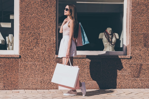 After day shopping. Side view of beautiful young woman carrying shopping bags and smiling while walking along the street with fashion store in the background