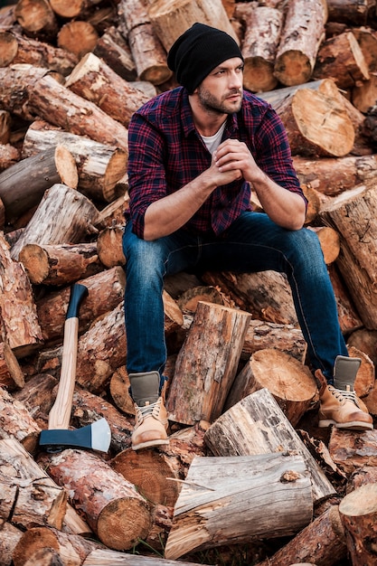 After hard physical work. Full length of thoughtful young forester keeping hands clasped and looking away while sitting on logs