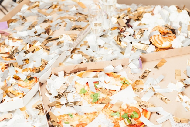 After party mess Closeup of pizza doughnut leftovers covered with shiny confetti Half empty champagne glasses