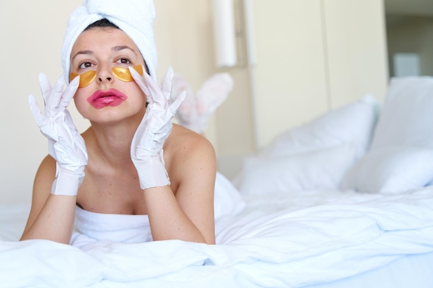 After a shower a girl wrapped in a towel uses cosmetic patches for the skin under the eyes lips and gloves to moisturize her hands and feet Cosmetic trends for body care at home