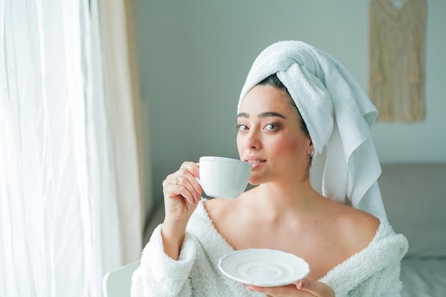 After a shower, a young woman in a bathrobe with a towel on her head drinks coffee and looks out the window