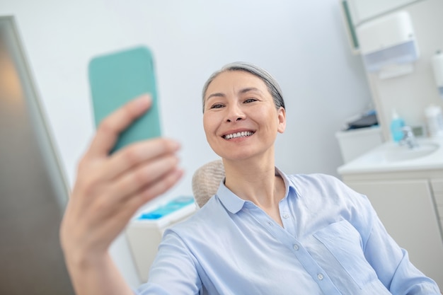 After teeth restoration. Smiling pretty mature woman with a smartphone at the dentists office