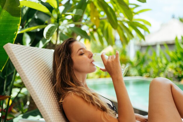 Sullo sfondo della piscina e del verde intorno ad essa siede una ragazza e beve da un bicchiere