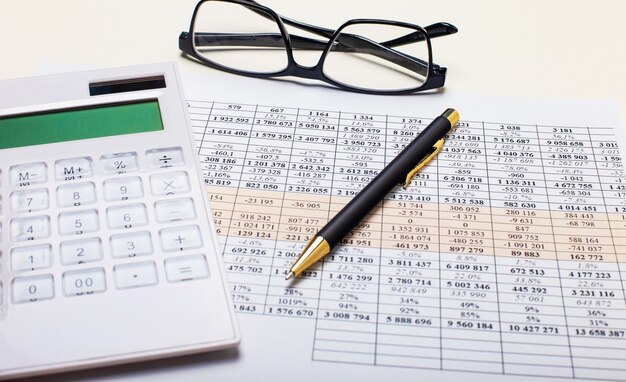 На фоне отчетов ручка, белый калькулятор и очки в черной оправе. Бизнес-концепция. На рабочем месте крупным планом