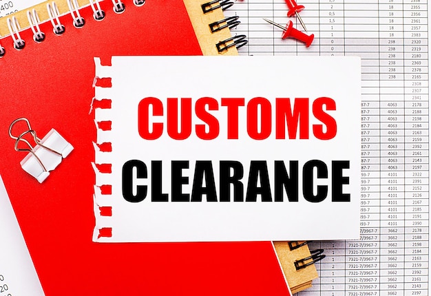 Against the background of the reports, there are red and brown notepads, a white paper clip, red buttons and a white sheet of paper with the text CUSTOMS CLEARANCE. Business concept