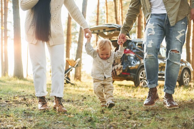 Photo against black automobile happy family of father mother and little daughter is in the forest