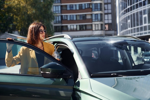 Against business building Young fashionable woman in burgundy colored coat at daytime with her car