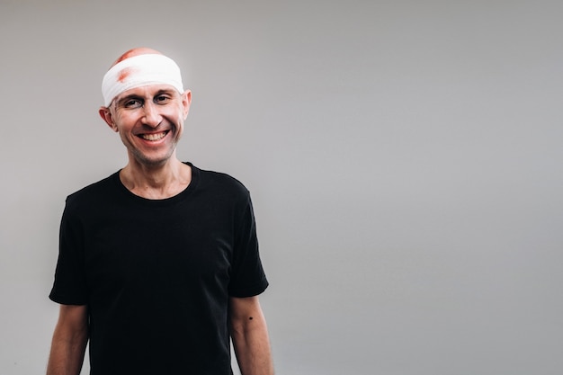 Against a gray background stands a battered and battered man in a black T-shirt with his head wrapped in a bandage.