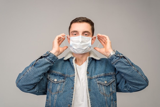 Against a light background, a man in a denim jacket puts on a medical mask