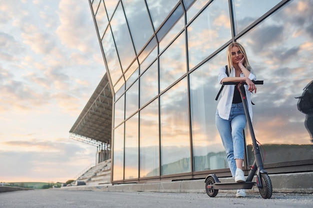 Against modern business building Beautiful blonde in casual clothes riding electric schooter outdoors at sunny daytime