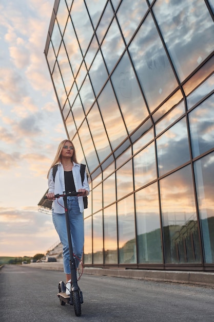 Against modern business building Beautiful blonde in casual clothes riding electric schooter outdoors at sunny daytime