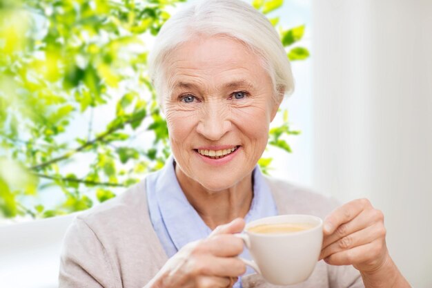 Age, drink and people concept - happy smiling senior woman with cup of coffee at home over window and green natural background