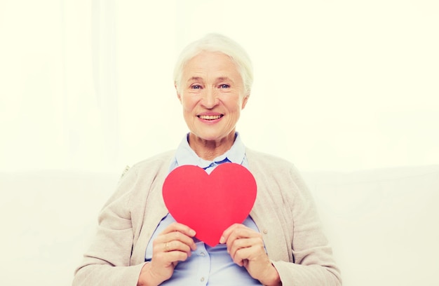 age, holidays, valentines day, love and people concept - happy smiling senior woman with red heart shape at home