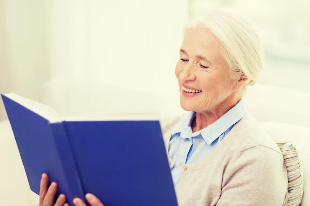 концепция возраста, отдыха и людей - счастливая улыбающаяся пожилая женщина читает книгу дома