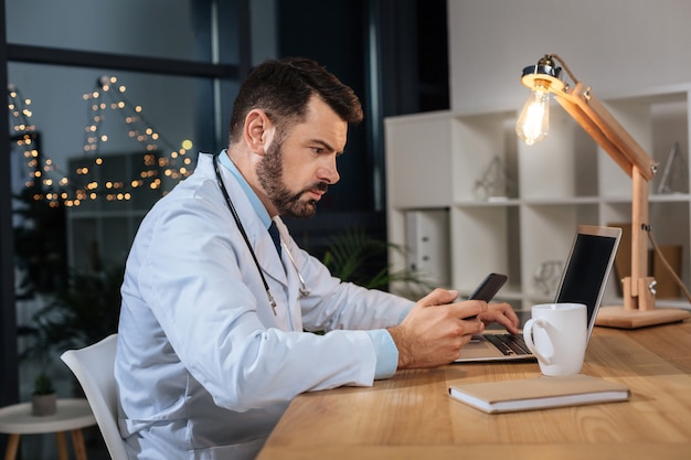 기술의 시대. 스마트 잘 생긴 남자 의사는 자신의 스마트 폰 화면을보고 그의 사무실에서 작업하는 동안 노트북을 사용
