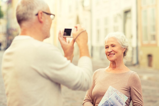 age, tourism, travel, technology and people concept - senior couple with map and camera photographing on street