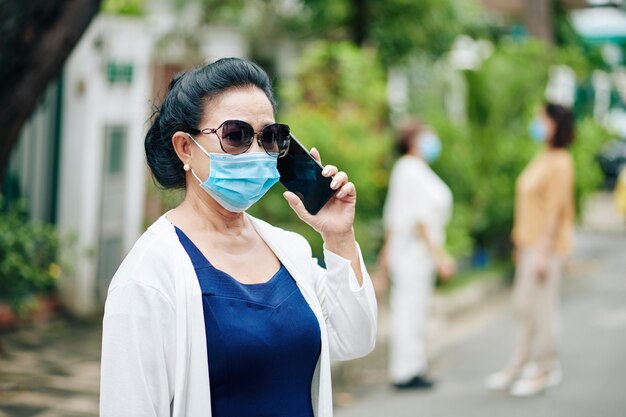 Aged Asian woman in sunglasses and medical mask walking outdoors and talking on phone