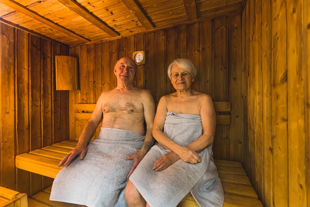 Aged caucasian couple with towels sitting in the wooden sauna and smiling medium full shot wellbeing