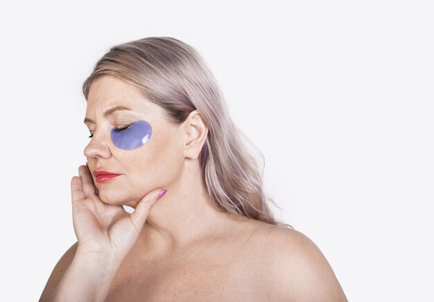 Aged caucasian woman with blonde hair and hydrogel eye patches posing with naked shoulders on a white  wall with free space