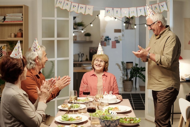 Aged cheerful woman in birthday cap looking at her friends clapping hands