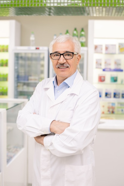 Aged drugstore worker in whitecoat and eyeglasses crossing his arms by chest while standing by workplace against displays with medical items