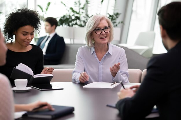 Aged female mentor conducting meeting with young professionals
mix race business people group listen to experienced leader or
coach while training young managers consulting with older
businesswoman