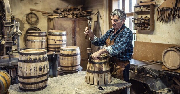 Aged man crafts a new vintage-looking wooden barrel using hammer to put the metal ring around the pieces of wood.