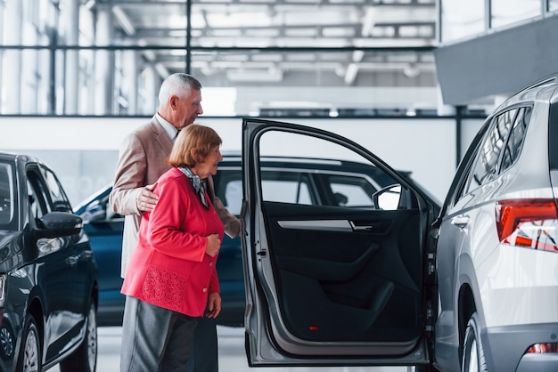 Aged man in formal wear supporting woman in choosing automobile.