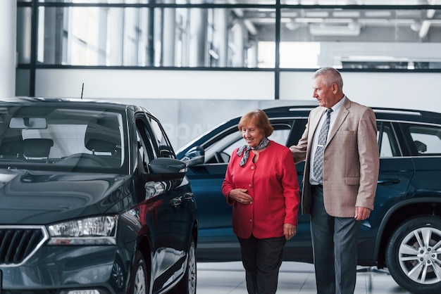 Aged man in formal wear supporting woman in choosing automobile.