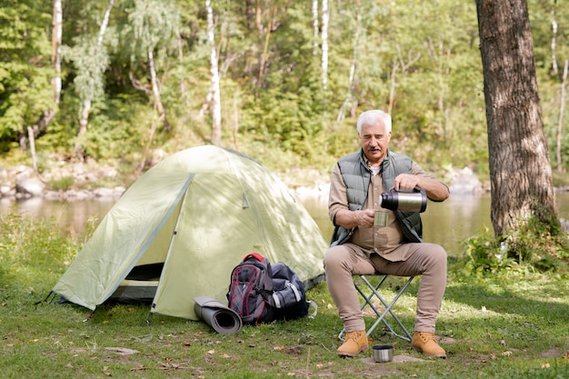 Aged man pouring hot tea or water into cup while sitting on small travel chair by tourist tent in the forest