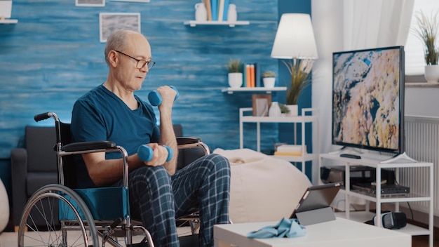 Aged man sitting in wheelchair and lifting weights while\
following video of workout exercise. senior person with physical\
disability using dumbbells and watching training lesson on\
tablet