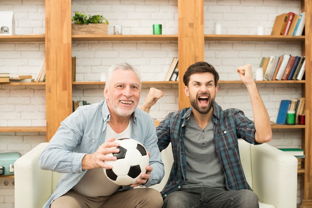 Aged man with ball and young crying guy watching TV on sofa