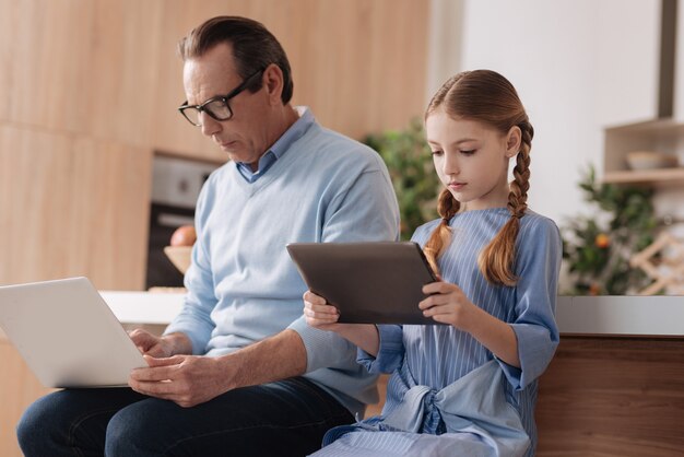 Aged motionless retired man sitting indoors and using gadgets with granddaughter while surfing the Internet and checking social media profiles