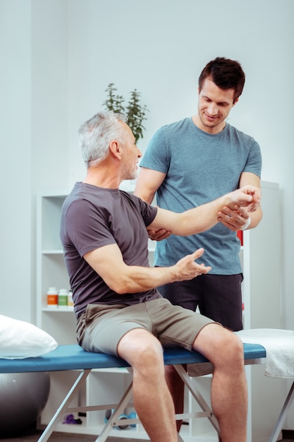 Aged patient. Nice senior man speaking with his nurse while sitting on the medical bed