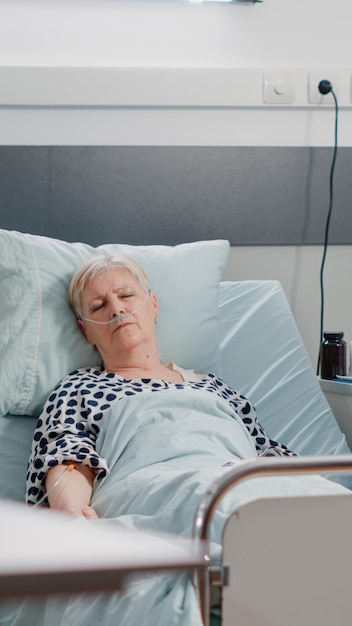 Aged patient with illness resting in hospital ward bed while
nurse looking at heart rate monitor for examination. sick woman
having nasal oxygen tube and iv drip bag for healthcare