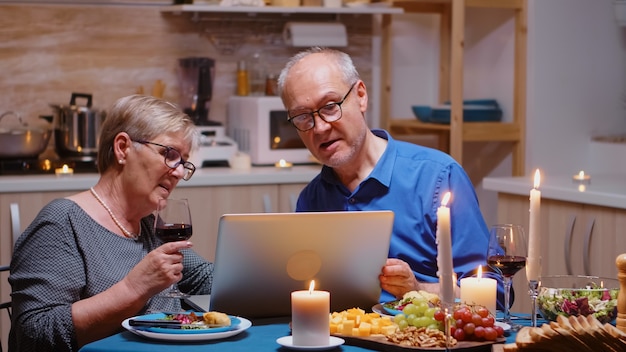 Aged, retired, couple shopping online during romanitic dinner using laptop. Old people sitting at the table, browsing, using the technoloty, internet, celebrating their anniversary in the dining room.