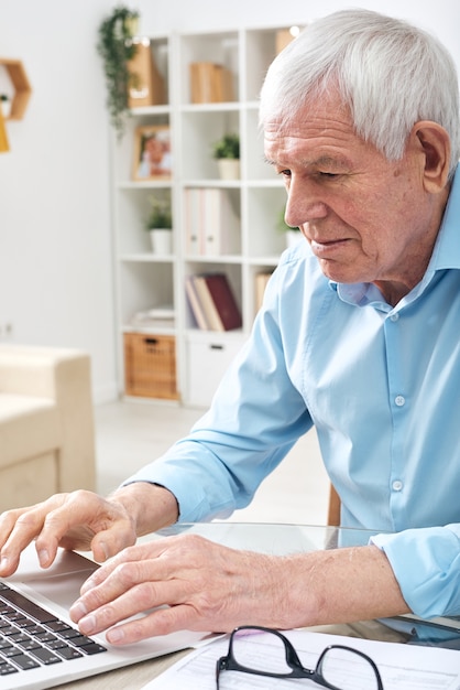 Aged retired man in blue shirt pressing keys of laptop keypad while surfing in the net for online data in home environment